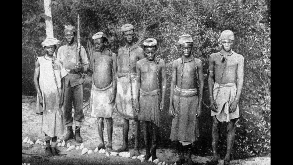 Historical photo of five enslaved or captive men in neck chains standing next to an armed guard